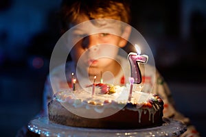 Happy blond little kid boy celebrating his birthday. Child blowing seven candles on homemade baked cake, indoor