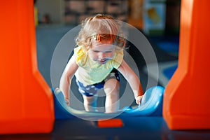 Happy blond little toddler girl having fun and sliding on indoor playground at daycare or nursery. Positive funny baby