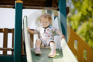 Happy blond little toddler girl having fun and sliding on outdoor playground. Positive funny baby child smiling.