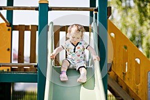 Happy blond little toddler girl having fun and sliding on outdoor playground. Positive funny baby child smiling. Summer