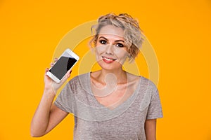 Happy blond woman in sweater showing blank smartphone screen and pointing on it while looking at the camera with open