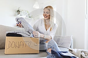 Happy blond young woman sit on couch stuck clothes in donation box at home, caring biracial female volunteer put apparel in carton