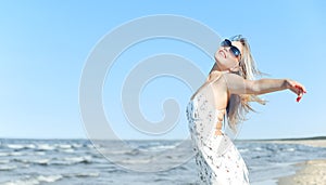 Happy blonde beautiful woman on the ocean beach standing in a white summer dress and sun glasses, open arms