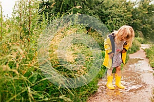 Happy blonde child girl in a yellow rubber boots and yellow raincoat staying in a puddle. Summer, childhood, holiday