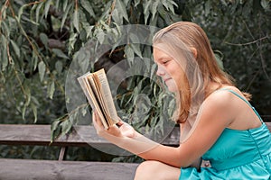 Happy blonde girl in a blue dress sitting on bench and reading book in park. Side view