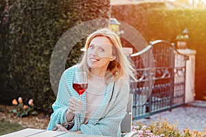Happy blonde girl enjoying red wine outdoors. Smiling attractive joyful woman in light blue knitted plaid smiling and looking at g