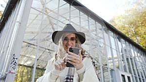 Happy blonde hair woman in sunglasses typing message in the smartphone on the park background.