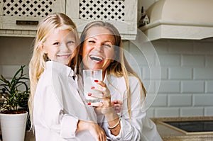 Happy blonde long hair mom and daughter drinking milk in bright kitchen, healthy lifestyle