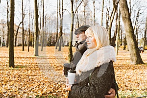 Happy blonde mature woman and handsome middle-aged brunette man walk in park and drink coffee from a reusable cup. A loving couple