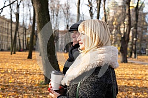 Happy blonde mature woman and handsome middle-aged brunette man walk in park and drink coffee from reusable cup. A loving couple