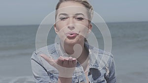 Happy blonde woman in denim jacket sending a kiss on lonely beach.