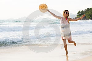 Happy blonde woman with hat in hand, enjoys her tropical vacation at beach, in Seychelles