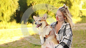 Happy blonde woman holding a Pembroke Welsh Corgi dog in her arms at sunset on a summer day while walking. Happy friends