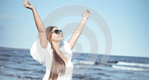 Happy blonde woman is on the ocean beach in a white dress and sunglasses, raising hands