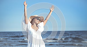 Happy blonde woman is on the ocean beach in a white dress and sunglasses, raising hands