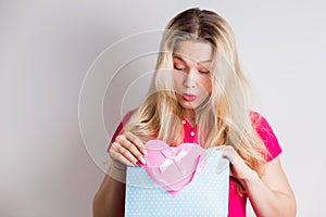 Happy blonde woman in a pink dress received a gift for a birthday or Valentine`s day. Cute girl opens blue package and smiles,