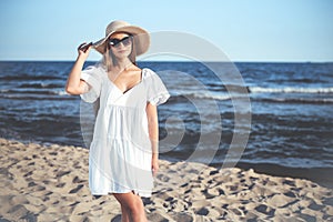 Happy blonde woman is posing on the ocean beach with sunglasses and a hat. Evening sun