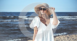 Happy blonde woman is posing on the ocean beach with sunglasses and a hat. Evening sun