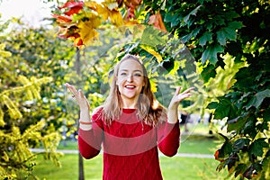 A happy blonde woman in a red sweater throws up bright autumn leaves. A happy smiling girl in the park. Seasonal Autumn concept