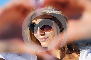 Happy blonde woman shows a heart symbol with her arms on the ocean beach, sunglasses, and a hat