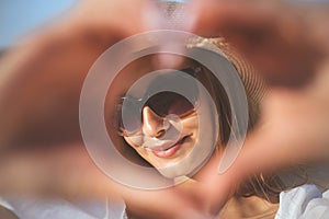 Happy blonde woman shows a heart symbol with her arms on the ocean beach, sunglasses, and a hat