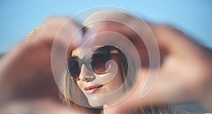 Happy blonde woman shows a heart symbol with her arms on the ocean beach, sunglasses, and a hat