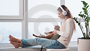 happy blonde woman sits on windowsill panoramic window headphones, listens cheerful driving music with cat animal dances