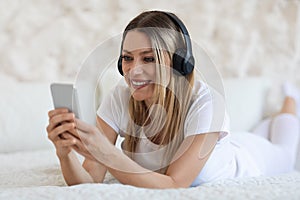 Happy blonde woman using cell phone and headset, bedroom interior