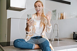 Happy blonde woman waving hand while using cellphone at home kitchen