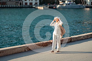 Happy blonde woman in a white suit and hat posing at the camera against the backdrop of the sea