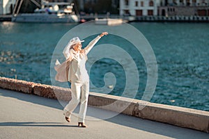 Happy blonde woman in a white suit and hat posing at the camera against the backdrop of the sea