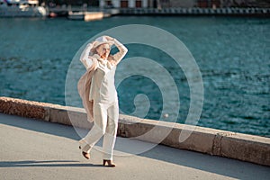 Happy blonde woman in a white suit and hat posing at the camera against the backdrop of the sea