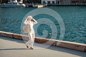 Happy blonde woman in a white suit and hat posing at the camera against the backdrop of the sea