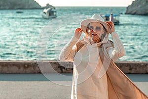 Happy blonde woman in a white suit and hat posing at the camera against the backdrop of the sea