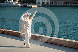 Happy blonde woman in a white suit and hat posing at the camera against the backdrop of the sea