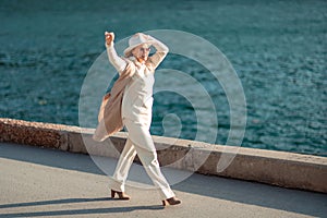 Happy blonde woman in a white suit and hat posing at the camera against the backdrop of the sea