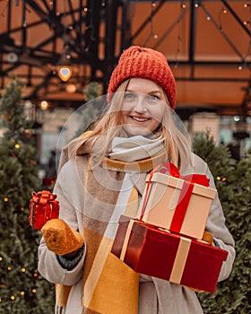 Happy blonde woman in winter coat holding a gift box at Christmas market. Attractive happy girl shopper hold gift boxes
