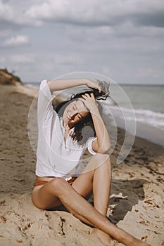 Happy boho girl in white shirt sitting on sunny beach. Carefree stylish woman smiling and relaxing on seashore. Summer vacation.