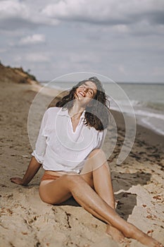 Happy boho girl in white shirt sitting on sunny beach. Carefree stylish woman smiling and relaxing on seashore. Summer vacation.
