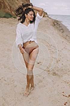 Happy boho girl in white shirt walking on sunny beach. Carefree stylish woman in swimsuit and shirt relaxing on seashore. Summer photo