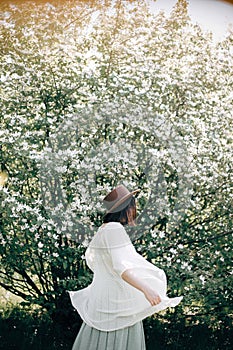 Happy boho woman in hat dancing in sunny light near blooming tree with white flowers in spring park. Stylish hipster girl enjoying