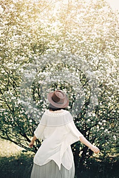 Happy boho woman in hat dancing in sunny light near blooming tree with white flowers in spring park. Stylish hipster girl enjoying