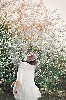 Happy boho woman in hat having fun in sunny light near white blooming tree in spring park. Stylish hipster girl enjoying spring