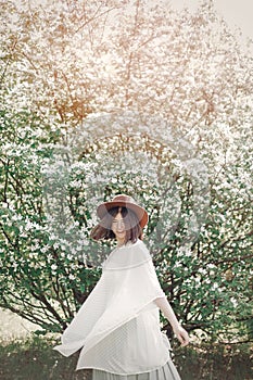 Happy boho woman in hat having fun in sunny light near white blooming tree in spring park. Stylish hipster girl enjoying spring