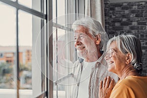 Happy bonding loving middle aged senior retired couple standing near window, looking in distance, recollecting good memories or