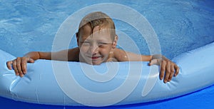 A happy boy with blond hair and blue eyes is swimming in an inflatable pool with clear blue clear water on a sunny summer day.