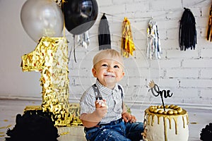 Happy boy blonde 1 year old on his birthday tries sweets. The photo zone with the first cake is decorated in black and gold.