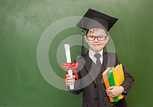 Happy boy with books and a diploma in graduation cap stands near a school board