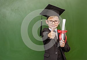 Happy boy with diploma in graduation cap stands near a school board and shows thumbs up gesture