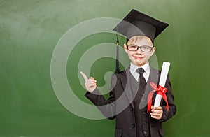 Happy boy with diploma in graduation hat points on empty green chalkboard